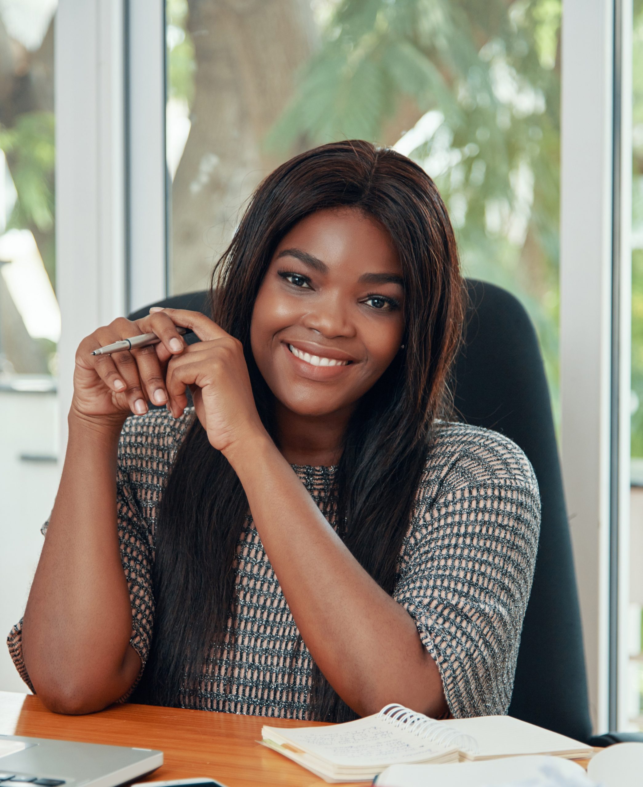 Happy adult black woman sitting at working desk in office smiling at camera adorably