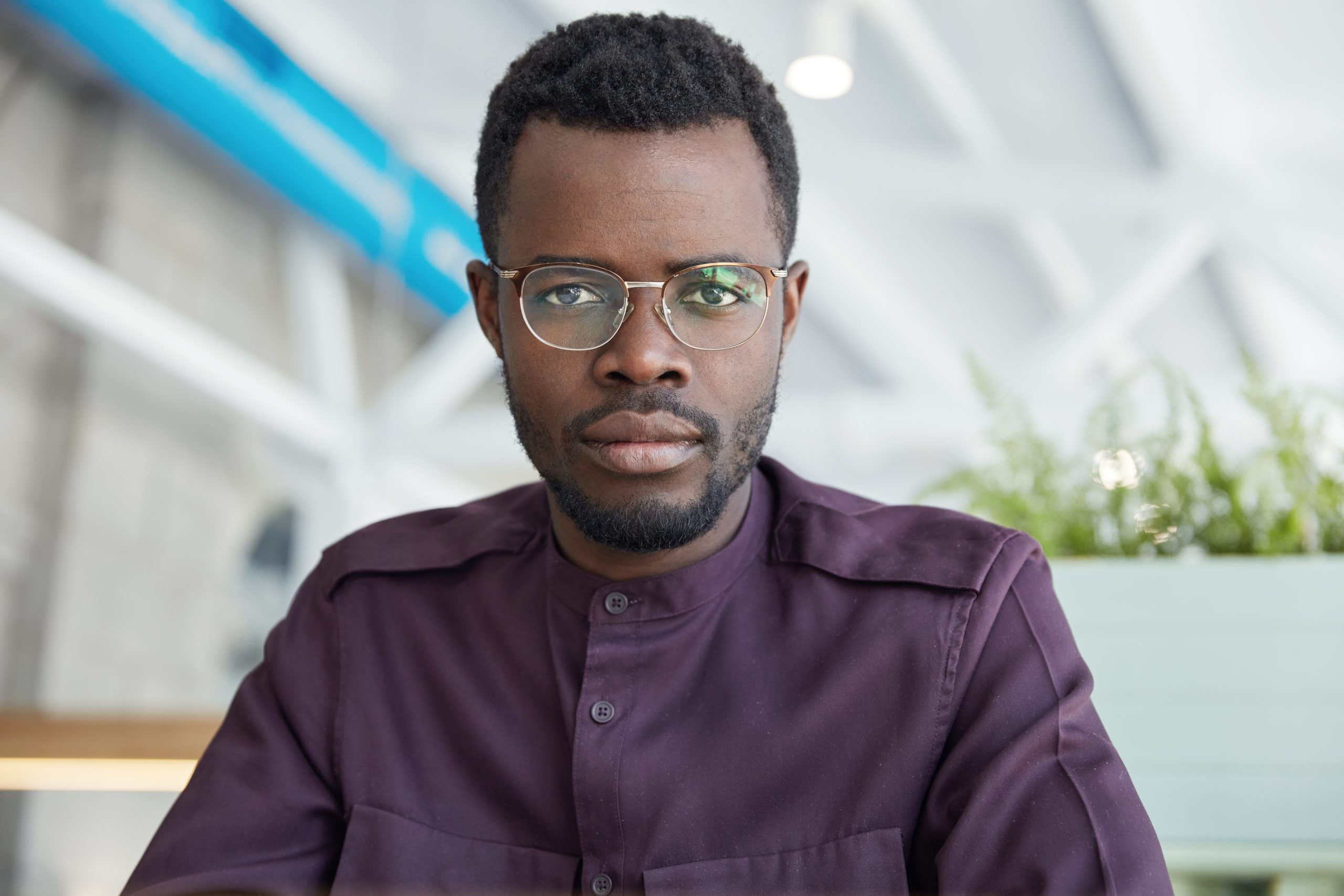 Portrait of serious confident male office worker in spectacles and formal shirt, with dark skin, poses in spacious cabinet, has formal meeting with business partners. People, ethnicity concept